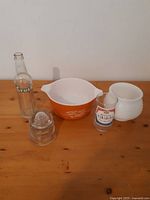 Full view of all glassware items on wooden table: Pyrex bowl, milk glass bowl, Orange Crush bottle, glass insulator, Canadian shot glass.