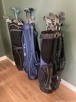 Three golf bags filled with various golf clubs standing against a wall on a hardwood floor.