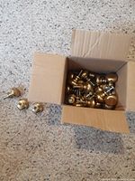 Box filled with approximately 30 vintage brass hardware knobs with screws attached, placed on carpeted floor. Two brass knobs outside the box for display.