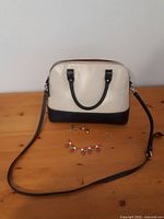 Handbag with cream upper body, black top handles and bottom trim, detachable black strap laid out on wooden surface with colorful charm necklace in front.