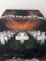 Photo of black Metallica flag with Master of Puppets album artwork showing crosses with puppet strings and sunset background.