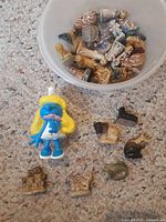 Plastic Smurfette surrounded by multiple small porcelain tea figurines including teapots, cups, and kettles inside a plastic container and on carpeted floor.