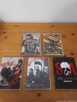 Five DVD cases from Sons of Anarchy Seasons 1 to 5 placed on a wooden surface. Covers show detailed artwork and series branding.