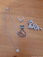 Four jewelry items including a Swarovski crystal pendant necklace, a sterling silver ring, a dragon brooch, and a cat brooch on a wooden surface.
