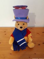 Winnie the Pooh plush wearing a red shirt, blue scarf with reflective stripes, and a tall purple hat with a red bow