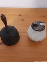 Side and top view of the metal bell and white glass table lighter on a wooden surface showing details and overall condition.