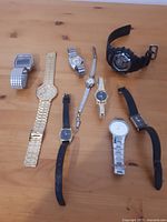 Photo showing nine various wristwatches on wooden surface including digital and analog styles with metal and leather/rubber bands.