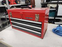 Red Craftsman metal toolbox sitting on a white surface showing front and top black handle with latches closed, three drawers visible