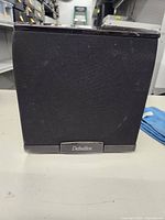 Front side view of the black cube-shaped subwoofer with foam grille and Definitive badge at the bottom center.