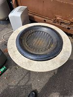 Top view of the firepit showing the black metal fire basin with grill grate inside, placed on beige stone tabletop with crackled texture