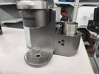 Front view of the silver Keurig coffee/latte/cappuccino machine showing the drip tray, control buttons, and a metal cup on the platform.