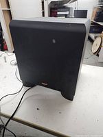 Side view of Klipsch subwoofer showing the fabric front and the power cord on a white surface.