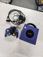 Photo showing purple GameCube console along with purple wired controller and black RFU adapter with cables.