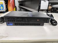 Sony PlayStation 2 console from front angled view showing disc tray, control buttons, and cords attached on sides on white surface.