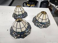 Three Tiffany style chandelier shades displayed on table, showing upper cream portion and blue leaf stained glass border