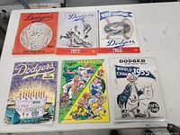 Six Los Angeles Dodgers yearbooks/magazines arranged on a table showing front covers with colorful vintage illustrations from years 1956 to 1971.