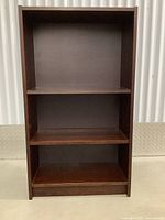 Front view of simple dark wood bookshelf with three shelves and loose backing, showing adjustable shelf peg holes.