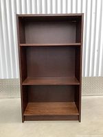 Front view of bookshelf showing three shelves, dark wood finish, and detached back panel needs reattachment.