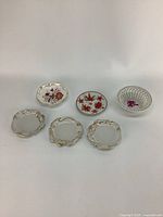 Five vintage small trinket dishes on white background including three ornate fine china pieces with gold detailing and floral motifs, one clear ribbed glass dish with purple flower, and one plate with orange leaf design.