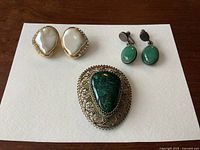 Photo showing three jewelry pieces: pearl-style gold-tone clip-on earrings with rhinestones, green stone sterling silver clip-on earrings, and a large vintage brooch with a green stone and gold-tone filigree setting.