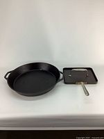 Two kitchenware items side by side on a white surface: round cast iron paella pan and rectangular panini press.