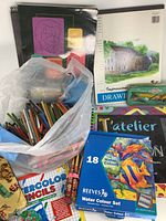 Photo showing Reeves 18-color watercolour set box with colorful fish artwork on front, surrounded by plastic bag filled with colored pencils and multiple sketch pads with black and artistic covers.