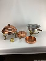 Photo showing set of vintage copper chafing dishes with a copper covered warming dish, a copper pan, a stainless steel lined chafing dish on brass legs, and a small brass fuel holder on white background.