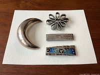 Four vintage sterling silver brooches and pins displayed on a white surface: crescent moon, flower shape, textured rectangle, and enameled rectangle with blue and multicolor design.