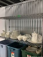 Minton fine china dinnerware arranged on metal shelves showing stacked plates, cups, and teapots with visible floral patterns and cream white finish.