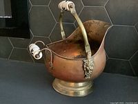 Front angled view showing the curved brass handle with porcelain grips and lion's head attachments, copper body and brass base with patina.