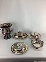 Front view of five silverplated serving pieces including a champagne bucket, two covered oval dishes, a round covered dish, and a small decorative bowl with cutouts.