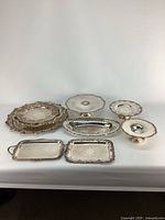 Nine vintage silverplated serving pieces arranged on a white surface showing variety of tray and bowl shapes with tarnishing and wear.