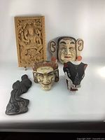 Front view of five wooden Asian decorative items on white background including four masks and one relief carving panel.