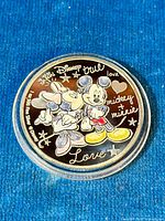 Obverse side of coin showing colorful Mickey and Minnie Mouse design with text and stars, inside a clear protective case on a blue background.