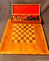 Open folding wooden chess board with inset checkered playing surface, wooden chess pieces in storage box with red felt lining inside the lid.