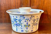 Front view of the covered casserole dish showing whimsical human and floral blue and green illustrations on white ceramic against a wooden background.