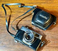 Photo shows Minolta Hi-Matic 7S camera with lens and attached carrying strap next to black leather case on wood surface.
