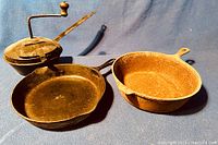 Three cast iron pieces: two frying pans and one metal coffee roaster on blue fabric