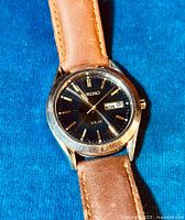 Front view of Seiko watch dial showing black face, gold-tone markers, and day/date display.