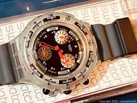 Close-up of the watch face showing black dial, multiple chronograph sub dials, world cities around bezel, orange and white hands, and translucent case with grey rubber strap.