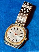 Close-up photo of the vintage Caravelle men’s watch on blue fabric background showing gold tone bezel, silver dial, black markers, red second hand, and stainless steel bracelet.