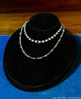 Two vintage sterling silver necklaces displayed on a black necklace stand: one short necklace with flower links and one longer necklace with eternity symbol links with blue cobalt wire wrapping.