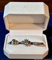 Set of three vintage sterling silver rings displayed inside a white padded ring box with hinged lid on blue background. Rings include a wave themed ring, a sun face ring, and a tree branch ring.