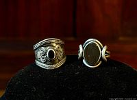 Two vintage sterling silver rings displayed on black velvet, one 'cigar band' style with spiral decorations and onyx stone, one with bezel-set oval onyx and double band rope design.