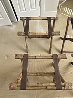 Two bamboo-style wooden luggage racks with brown fabric straps on beige carpet, arranged upright