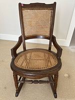 Front view of the antique wooden rocking chair showing the cane seat and backrest with wooden frame and armrests.