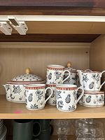 Ceramic kitchenware with floral and pineapple designs including mugs, covered pot, and small creamer.