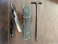 Photo showing the open straight razor, original storage case and opticians hammer placed on wooden surface