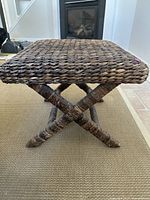 Side view of the dark brown woven wicker ottoman showing crossed legs and woven seat structure.