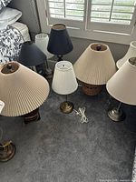 Eight lamps grouped on a carpeted floor near window with varied styles and colors; mix of beige, brown, white, and metallic shades and bases visible